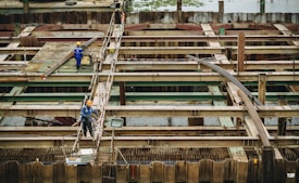 Workers in protective gear are seen on a construction site characterized by metal beams, scaffolding, and wooden supports. They appear to be engaged in construction tasks on a large infrastructure project, possibly a bridge or a similar structure.