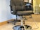 A black leather salon chair with a chrome base is positioned on a tiled floor in a well-lit room. Behind the chair, there is a trolley with wheels and bottles, indicating a salon or barbershop setting. The wall features a subtle light-colored paint.