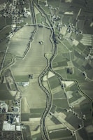 An aerial view of an agricultural landscape featuring numerous fields of varying sizes and shapes, bordered by a winding river. Small clusters of buildings or houses are scattered throughout the area, along with a network of roads connecting them.