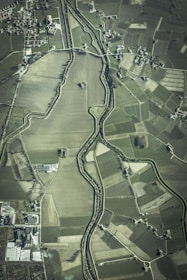 An aerial view of an agricultural landscape featuring numerous fields of varying sizes and shapes, bordered by a winding river. Small clusters of buildings or houses are scattered throughout the area, along with a network of roads connecting them.