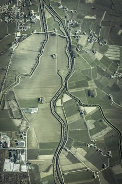 An aerial view of an agricultural landscape featuring numerous fields of varying sizes and shapes, bordered by a winding river. Small clusters of buildings or houses are scattered throughout the area, along with a network of roads connecting them.