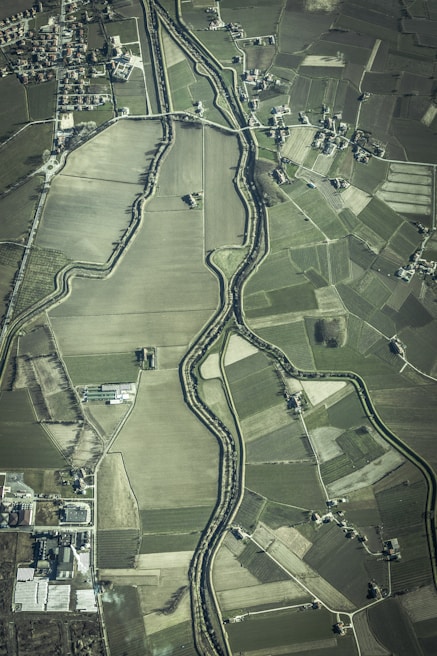 An aerial view of an agricultural landscape featuring numerous fields of varying sizes and shapes, bordered by a winding river. Small clusters of buildings or houses are scattered throughout the area, along with a network of roads connecting them.