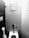 A monochrome photograph displaying a public restroom stall featuring a toilet with a flushometer, a wall-mounted toilet paper holder with a visible roll, and a small square mirror mounted on a tiled wall.