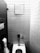 A monochrome photograph displaying a public restroom stall featuring a toilet with a flushometer, a wall-mounted toilet paper holder with a visible roll, and a small square mirror mounted on a tiled wall.