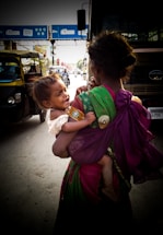 A woman dressed in a colorful sari carries a crying child in a busy street setting. The scene includes vehicles like auto rickshaws and a bus, and appears to be taken in an urban area with visible traffic and overhead signage.