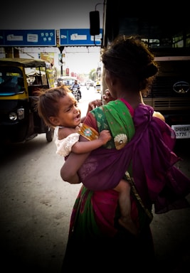 A woman dressed in a colorful sari carries a crying child in a busy street setting. The scene includes vehicles like auto rickshaws and a bus, and appears to be taken in an urban area with visible traffic and overhead signage.