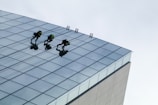 Three people are engaged in window cleaning or maintenance on the side of a tall, modern glass building. They are using ropes and harnesses for safety as they perform their tasks. The building features a grid of reflective glass panels.