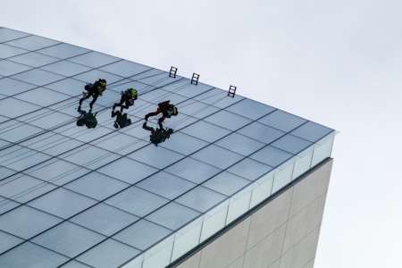 Three people are engaged in window cleaning or maintenance on the side of a tall, modern glass building. They are using ropes and harnesses for safety as they perform their tasks. The building features a grid of reflective glass panels.