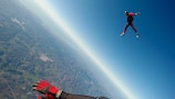 A skydiver dressed in a red and black suit is freefalling above a vast landscape with fields and hills visible below. The sky is clear and blue, with the horizon slightly curved, suggesting a high altitude. A second person's gloved hand is visible in the lower left corner, indicating the presence of another skydiver nearby.