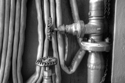 A monochrome image featuring a coiled fire hose alongside a metallic pipeline and valve. The valve has a round handle and appears to be an integral part of an industrial or mechanical setting.