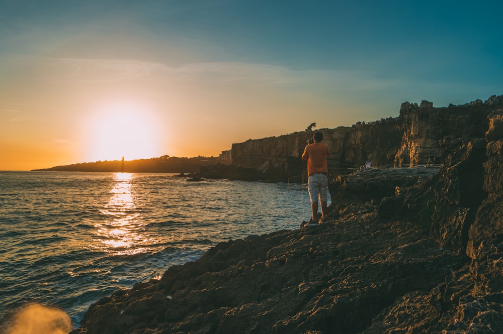 man standing on cliff beside body of water at daytime