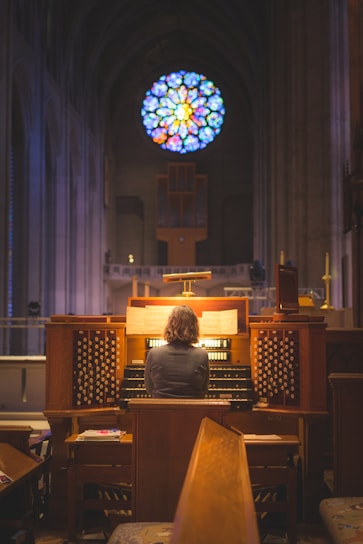 A person sits at a large wooden pipe organ in a church, bathed in the glow of surrounding lights. The organ console is intricate with numerous keys and stops. Above, a vibrant circular stained glass window casts colorful patterns.