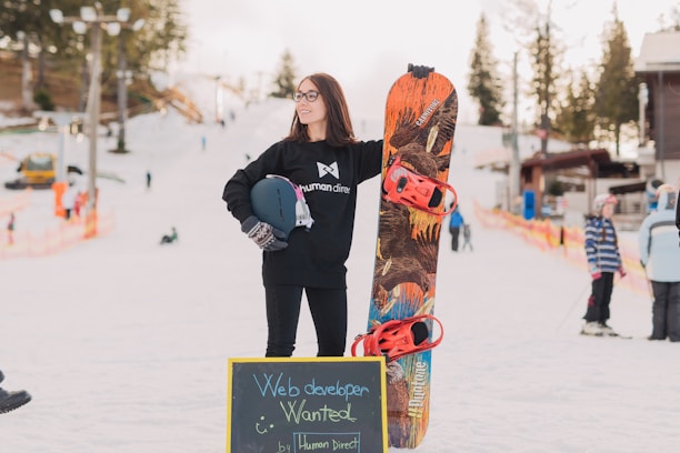A person stands confidently on a snowy slope holding a snowboard with colorful red bindings. They are wearing a dark sweatshirt and gloves, with a helmet tucked under one arm. In the background, other people are skiing or snowboarding. A chalkboard sign placed in front reads 'Web developer wanted'. The environment features trees and ski resort buildings, with a slightly cloudy sky.
