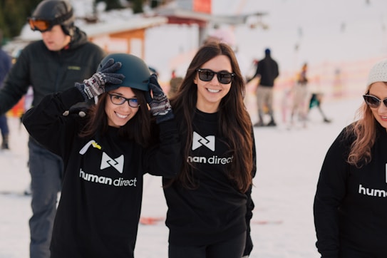 A group of people are outdoors in a snowy environment wearing black 'human direct' hoodies. One person is putting on a helmet, while the others are smiling and wearing sunglasses. There are others in the background walking or skiing in the snow.