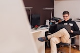A person sits comfortably in a chair, smiling while working on a laptop in a cozy office space. The desk nearby is organized with a computer monitor, a lamp, and a few office supplies. A small potted plant adds a touch of greenery to the setting.