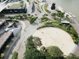 An aerial view of a complex featuring modern architecture, with curved pathways and well-maintained greenery. There's a sandy area that appears to be a volleyball court, surrounded by trees and walkways. A body of water is seen in the background, adjacent to a series of docks or boardwalks. The design is geometrical and organized, with a focus on aesthetics and greenery.