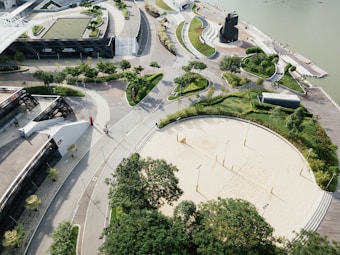 An aerial view of a complex featuring modern architecture, with curved pathways and well-maintained greenery. There's a sandy area that appears to be a volleyball court, surrounded by trees and walkways. A body of water is seen in the background, adjacent to a series of docks or boardwalks. The design is geometrical and organized, with a focus on aesthetics and greenery.