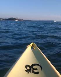 The tip of a yellow kayak or canoe with a rope attached, floating on a body of water. The water appears vast and slightly rippled under a clear blue sky. In the distance, there are land formations with some vegetation and a couple of boats.