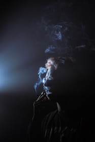 A person is seen in profile view holding a cigarette to their lips, with smoke swirling around their head. Light illuminates the smoke, creating a moody and mysterious atmosphere.