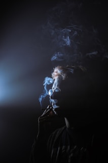 A person is seen in profile view holding a cigarette to their lips, with smoke swirling around their head. Light illuminates the smoke, creating a moody and mysterious atmosphere.