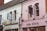 A quaint street scene featuring two distinct storefronts. One building has white painted brick with black signage for 'The Suite' and 'Intimate Lingerie'. Adjacent to it is a pink building with signage for 'om diva' and 'atelier 27'. Both structures have arched windows and decorative facades, capturing an eclectic and charming style.