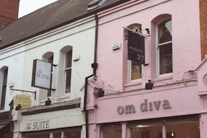 A quaint street scene featuring two distinct storefronts. One building has white painted brick with black signage for 'The Suite' and 'Intimate Lingerie'. Adjacent to it is a pink building with signage for 'om diva' and 'atelier 27'. Both structures have arched windows and decorative facades, capturing an eclectic and charming style.