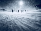 A snow-covered landscape with a group of skiers moving through a strong, blizzard-like snowstorm. The sky is overcast, and the sun is barely visible through the dense flurry of snow. Shadows of structures can be seen in the background, suggesting a mountainous or ski resort area.