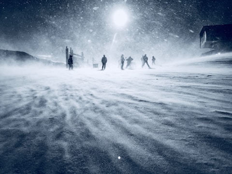 A snow-covered landscape with a group of skiers moving through a strong, blizzard-like snowstorm. The sky is overcast, and the sun is barely visible through the dense flurry of snow. Shadows of structures can be seen in the background, suggesting a mountainous or ski resort area.