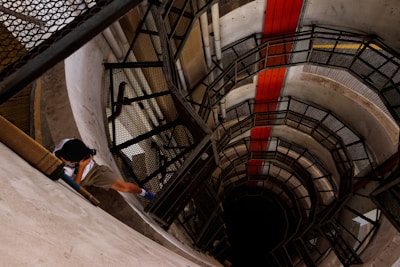 A person is photographing the interior of a circular, multi-story building with a spiral staircase. The structure is primarily made of concrete and metal, with a red stripe running vertically along the wall. Shadows play across the surfaces, creating a sense of depth.