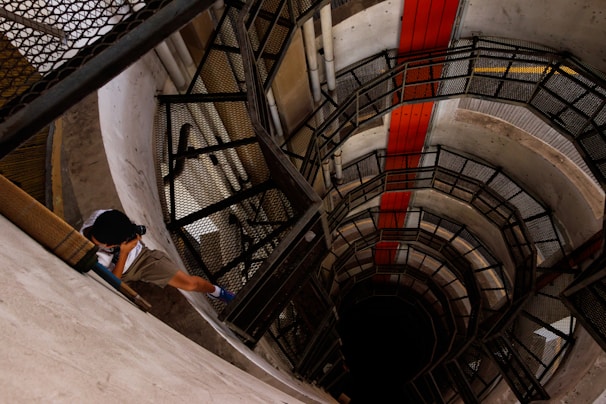 A person is photographing the interior of a circular, multi-story building with a spiral staircase. The structure is primarily made of concrete and metal, with a red stripe running vertically along the wall. Shadows play across the surfaces, creating a sense of depth.