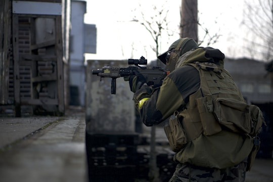 A person in tactical gear is holding a rifle and appears to be in a focused stance. The setting looks like an urban environment with industrial elements such as metal structures and concrete. The person's gear is detailed with a camouflage pattern and includes a protective helmet and vest.