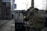 A person in tactical gear is holding a rifle and appears to be in a focused stance. The setting looks like an urban environment with industrial elements such as metal structures and concrete. The person's gear is detailed with a camouflage pattern and includes a protective helmet and vest.