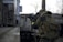 A person in tactical gear is holding a rifle and appears to be in a focused stance. The setting looks like an urban environment with industrial elements such as metal structures and concrete. The person's gear is detailed with a camouflage pattern and includes a protective helmet and vest.