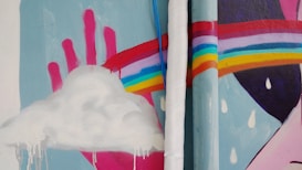 A colorful mural featuring a white, fluffy cloud with drips beneath it. A large pink hand is visible in the background, with fingers pointing upwards. A vibrant rainbow arches across the image, with raindrops scattered throughout. Two cylindrical pipes are positioned vertically in the center of the mural, intersecting with the rainbow.