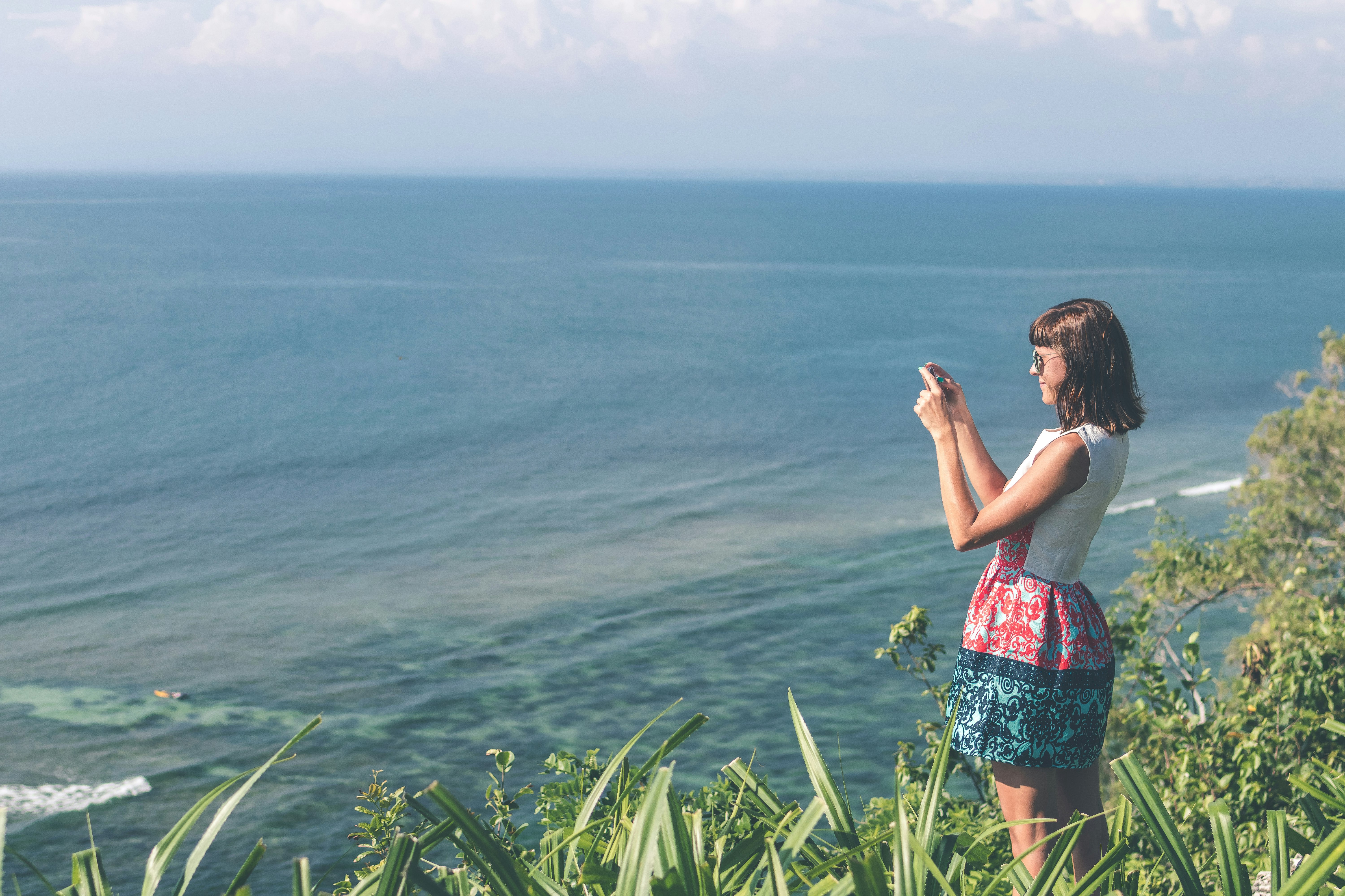 woman taking photo of ocean atop a cliff during day time