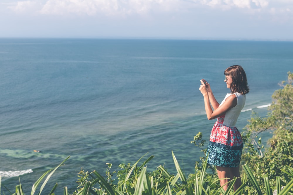 woman taking photo of ocean atop a cliff during day time