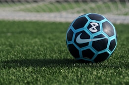 A blue and black soccer ball with a white Nike logo, resting on a green grass field. The background shows the blurred lines of a soccer goal net.