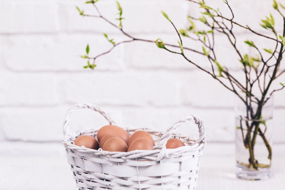 A white wicker basket containing brown eggs is placed on a surface. In the background, a clear glass vase holds a branch with young green leaves. The setting appears to be minimalist with a white brick wall as the backdrop.