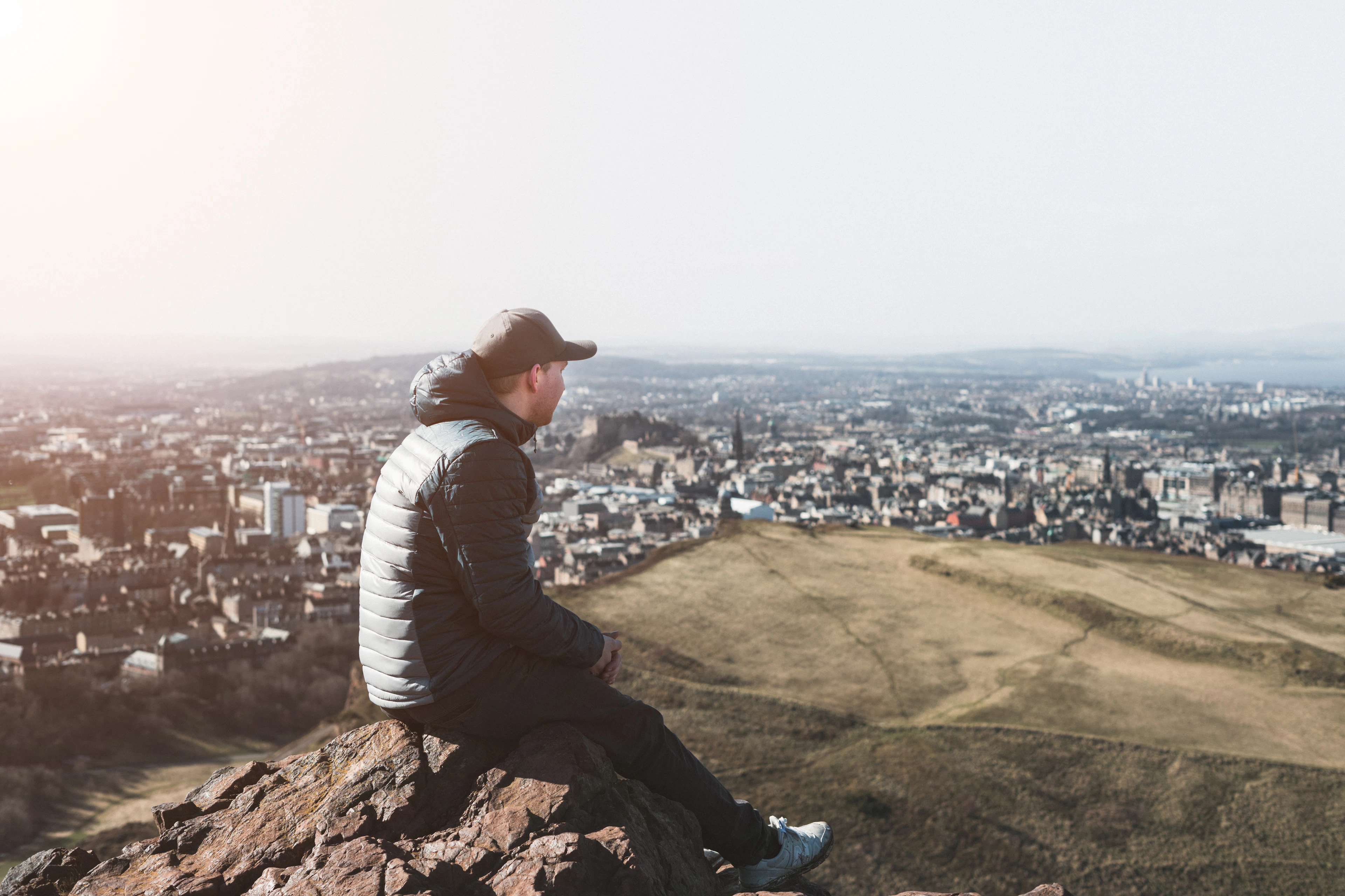 A walker sitting on the rocky summit of Arthur's Seat, looking out over the rooftops of Edinburgh in hazy afternoon light
