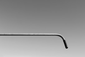 A minimalist image featuring a single metal pipe with a slight bend towards the right end. The background is a smooth gradient transitioning from dark gray at the top to light gray at the bottom, emphasizing the simplistic and abstract nature of the composition.