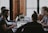 A group of people are seated at a table in a workspace setting, each focused on their laptops. The room is softly lit, giving it a cozy and productive atmosphere. One person is wearing headphones, likely concentrating on their task. A small plant is placed on the table, adding a touch of greenery.