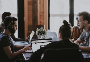 A group of people are seated at a table in a workspace setting, each focused on their laptops. The room is softly lit, giving it a cozy and productive atmosphere. One person is wearing headphones, likely concentrating on their task. A small plant is placed on the table, adding a touch of greenery.