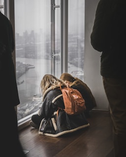Two individuals sit close together on a wooden floor in a high-rise building, leaning against a large window with a view of a foggy cityscape and river. One person has a brown backpack. A dark-clothed figure stands nearby, partially visible.