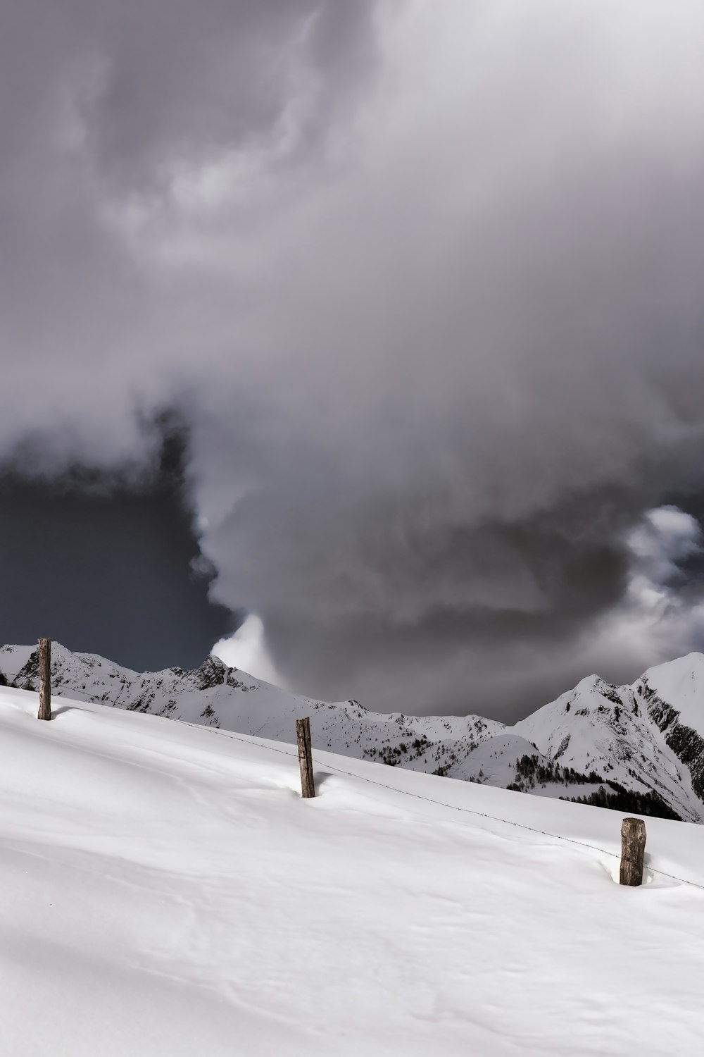 brown wooden post and barbed wire fence on snow surface overlooking mountains under grey cloudy sky