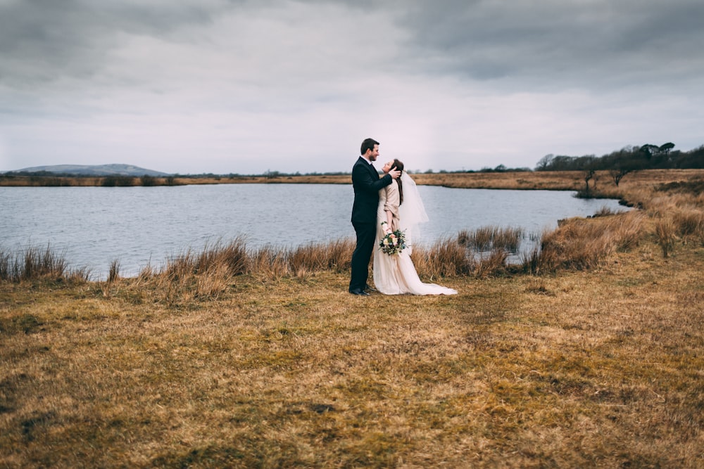 Photographie de mariée et de marié sur l’herbe brune près du plan d’eau pendant la journée