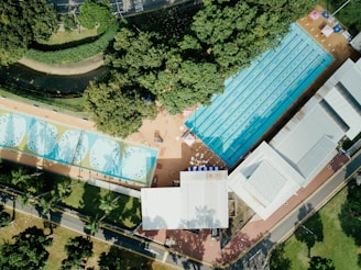 An aerial view of a swimming facility surrounded by greenery. The image features a large rectangular swimming pool with lanes, adjacent to smaller circular pools. There is a paved area with seating and umbrellas nearby, and various buildings along the perimeter.