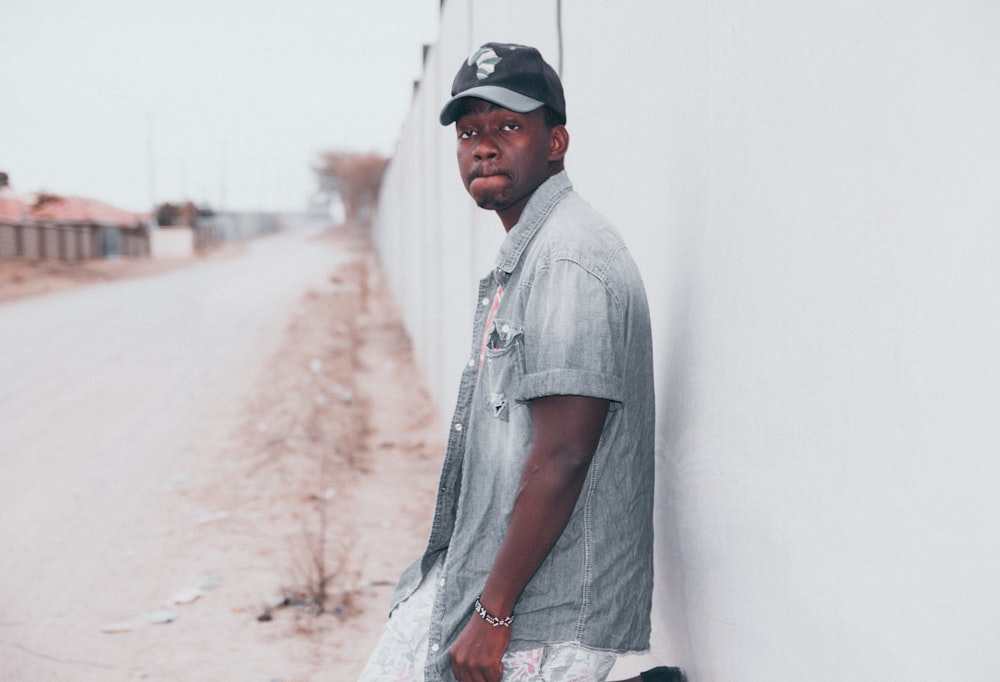 man in gray shirt and black cap leaning on white wall