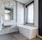 A modern bathroom features a sleek, rectangular bathtub positioned next to a window with blinds. The walls are adorned with textured tiles, and a rectangular mirror hangs above a white sink. A soft bath mat lies on the floor, and a towel is neatly placed on a wall-mounted rack.