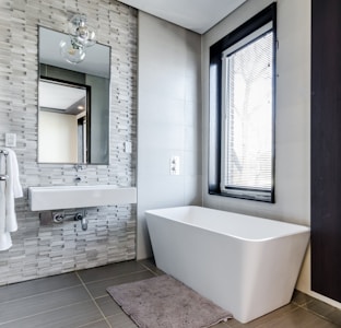 A modern bathroom features a sleek, rectangular bathtub positioned next to a window with blinds. The walls are adorned with textured tiles, and a rectangular mirror hangs above a white sink. A soft bath mat lies on the floor, and a towel is neatly placed on a wall-mounted rack.