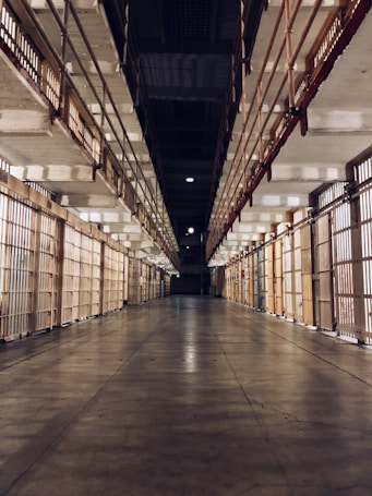 A long, narrow prison corridor flanked by barred cells on both sides, illuminated by overhead lights casting a pattern of shadows on the concrete floor.